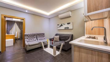 Ein modernes Wohnzimmer mit zwei Sofas, einem Glastisch, einer Küchenzeile mit Spüle und Armatur sowie einer Wand-Klimaanlage.