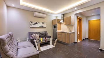 A modern living room with a sofa and a glass coffee table, a kitchen with wooden cabinets, and a wooden door.