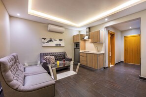A modern living room with a sofa and a glass coffee table, a kitchen with wooden cabinets, and a wooden door.