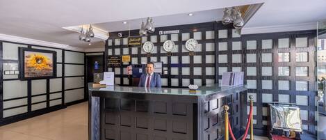 A hotel reception desk with a modern design, featuring a glass partition, a clock, and a sign indicating the reception area.
