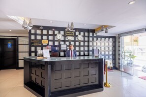 Un mostrador de recepción con un reloj, un cartel que indica habitación accesible, y una zona de recepción con una mampara de vidrio.