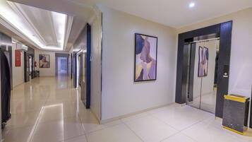 A hotel corridor with a modern design, featuring a long hallway, a wall-mounted abstract painting, and a glass elevator.