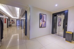 A hotel corridor with a modern design, featuring a long hallway, a wall-mounted abstract painting, and a glass elevator.