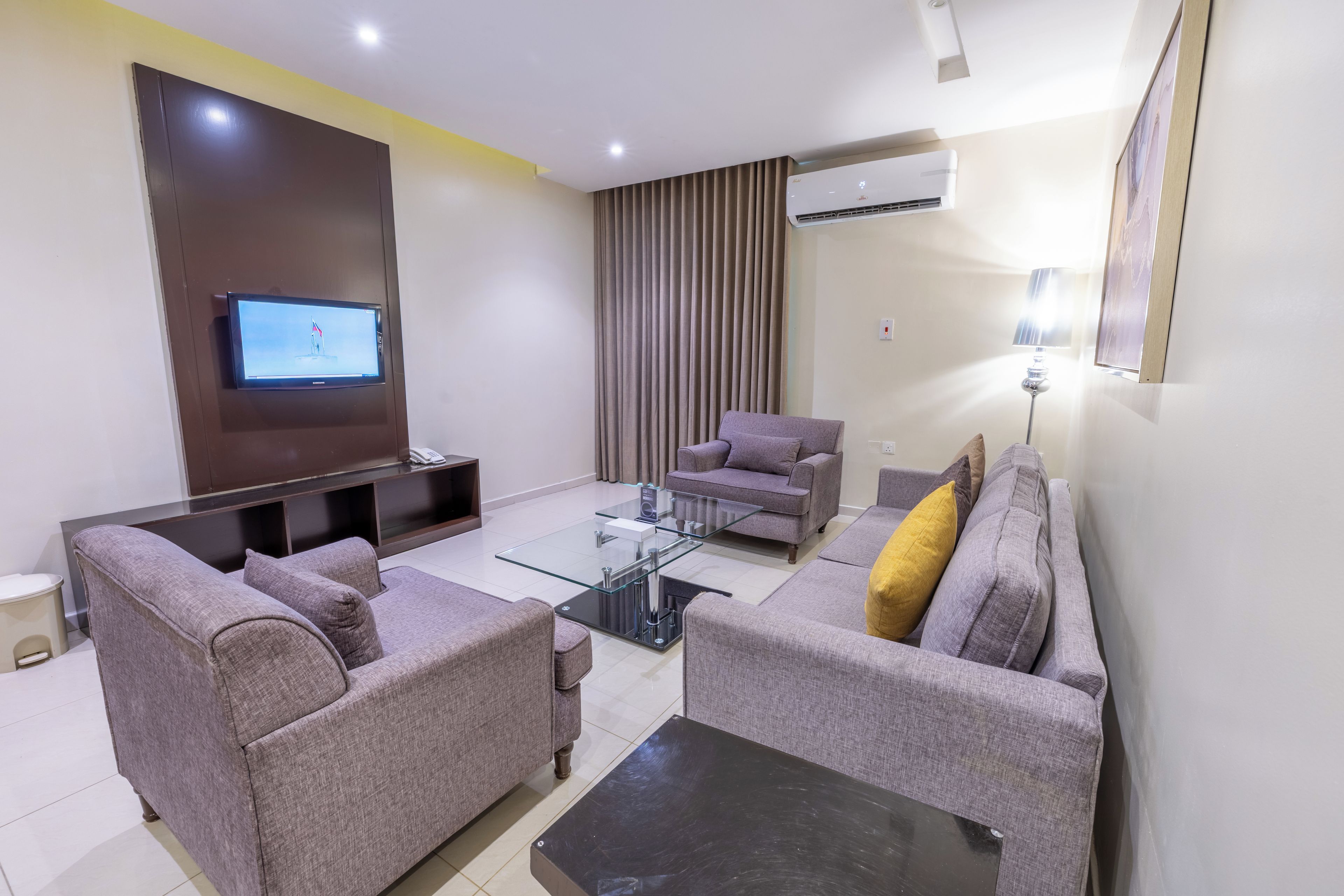 A modern living room with a flat-screen TV, two grey sofas, a glass coffee table, and an air conditioning unit.