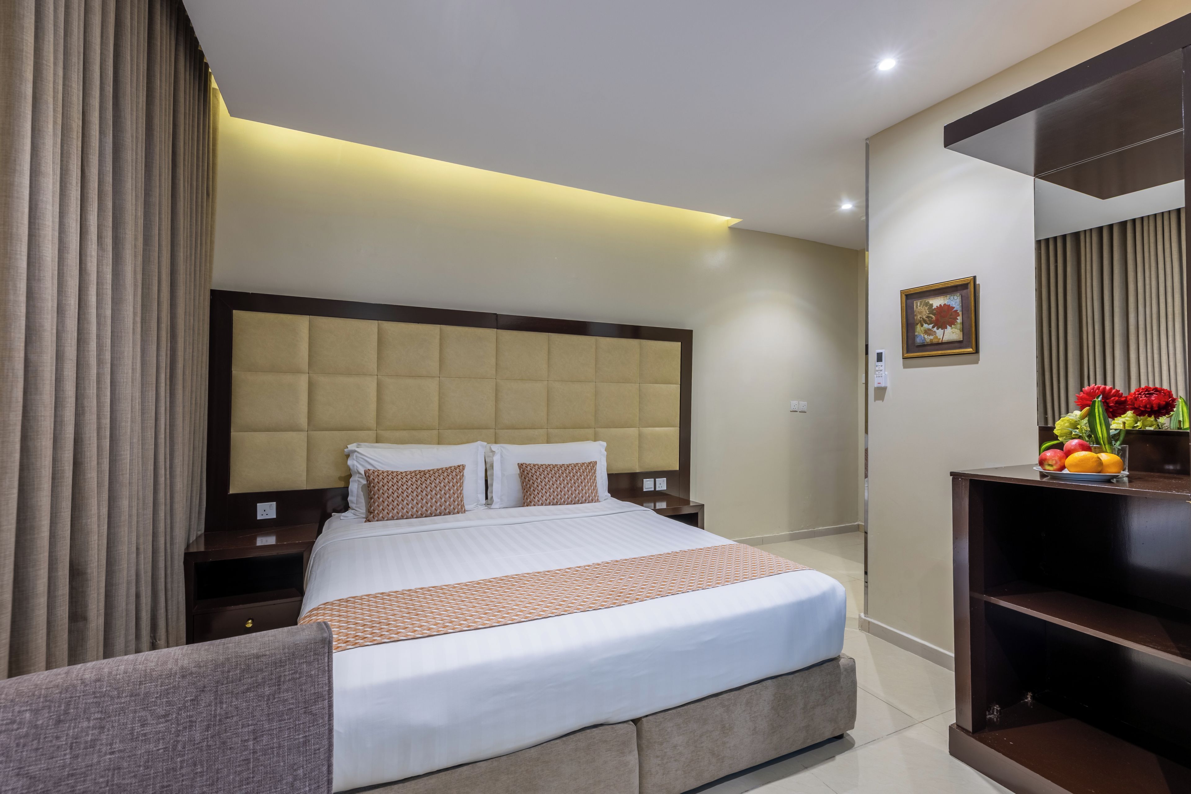 Een hotelkamer met een groot bed, een nachtkastje, een wandtelevisie en een klein plankje met fruit en bloemen.