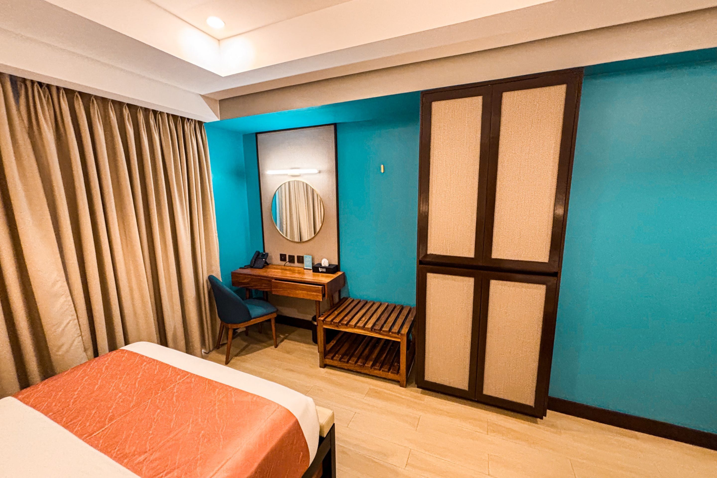 A hotel room with a bed, a desk, a chair, a small stool, a mirror, and a wardrobe.