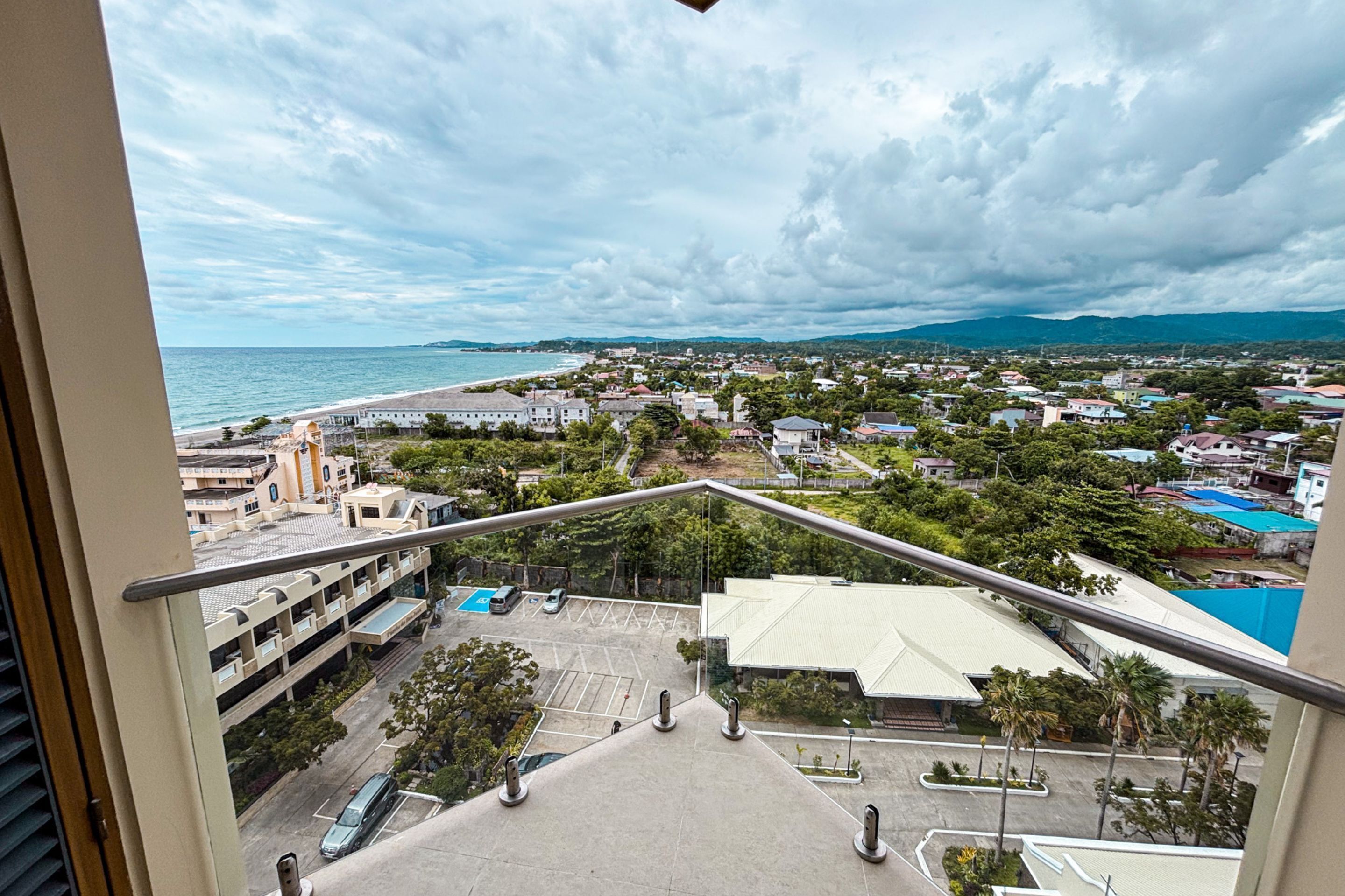 A balcony with a view of a coastal town, a parking area, and a building with a flat roof.
