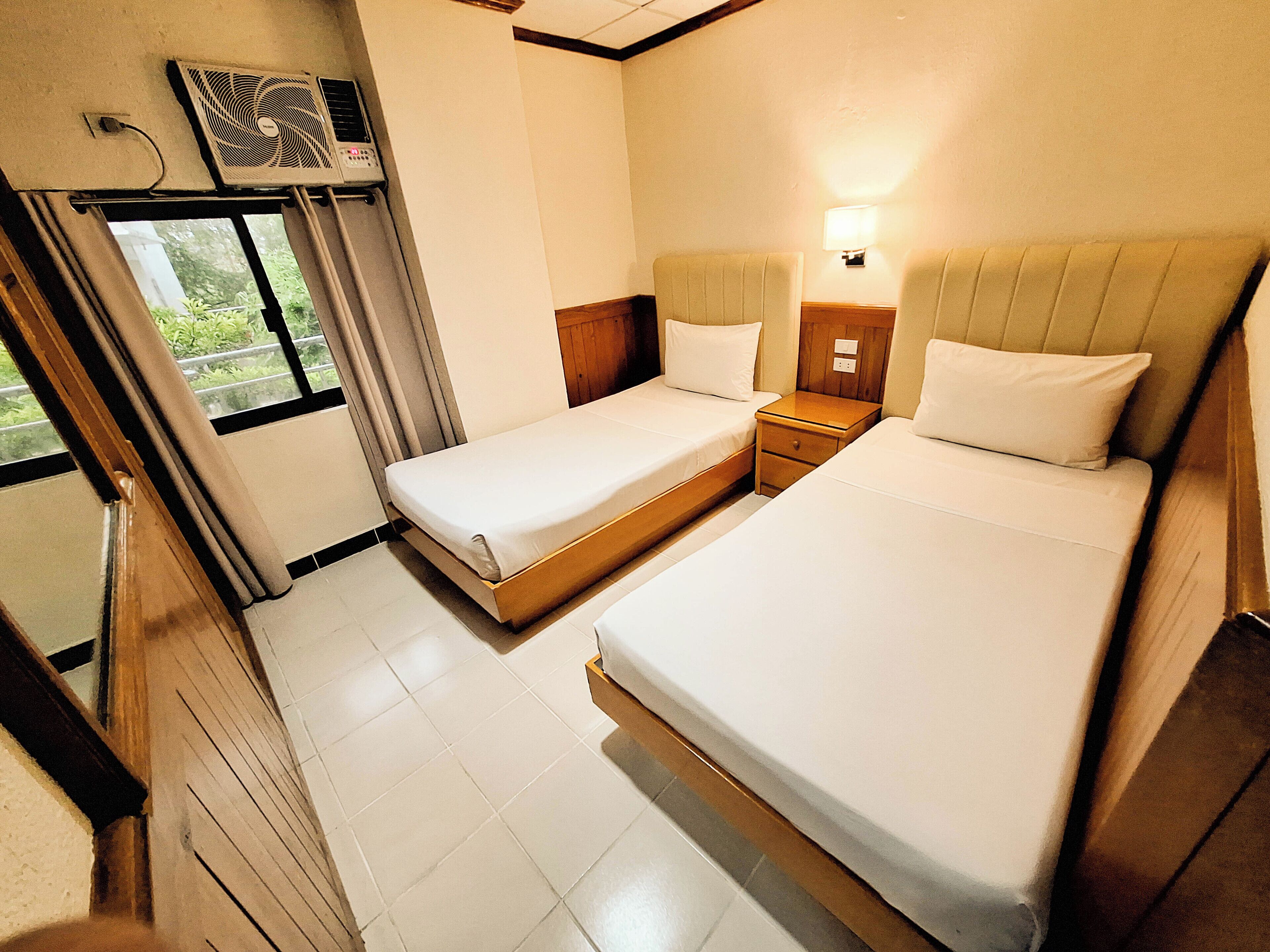 Habitación de hotel con dos camas, una mesita de noche pequeña, cortinas en una ventana y un aire acondicionado de pared.