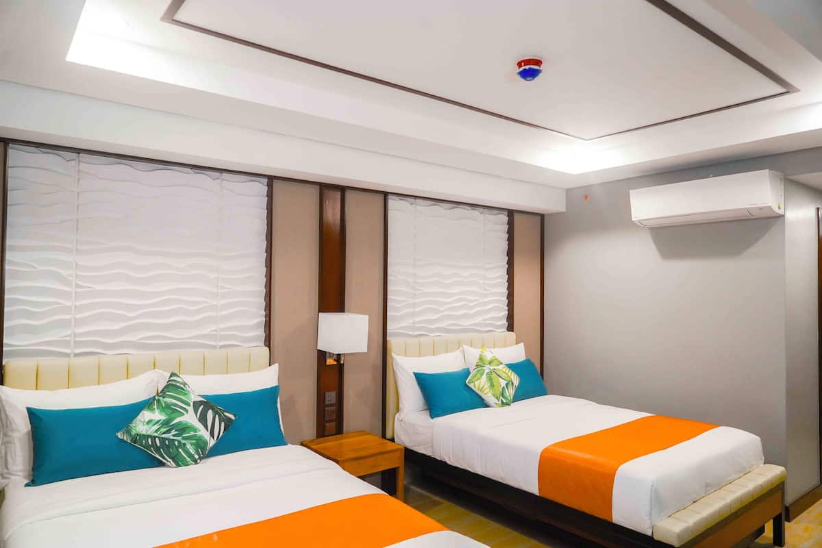 Ett hotellrum med två sängar, båda med vita sängkläder och orange detaljer, ett nattduksbord i trä, en väggmonterad luftkonditioneringsanläggning och en taklampa.