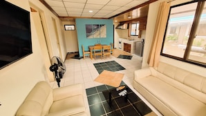 A living room with a sofa, a coffee table, and a TV. There is a dining area with a table and chairs, and a kitchen with a sink and cabinets.