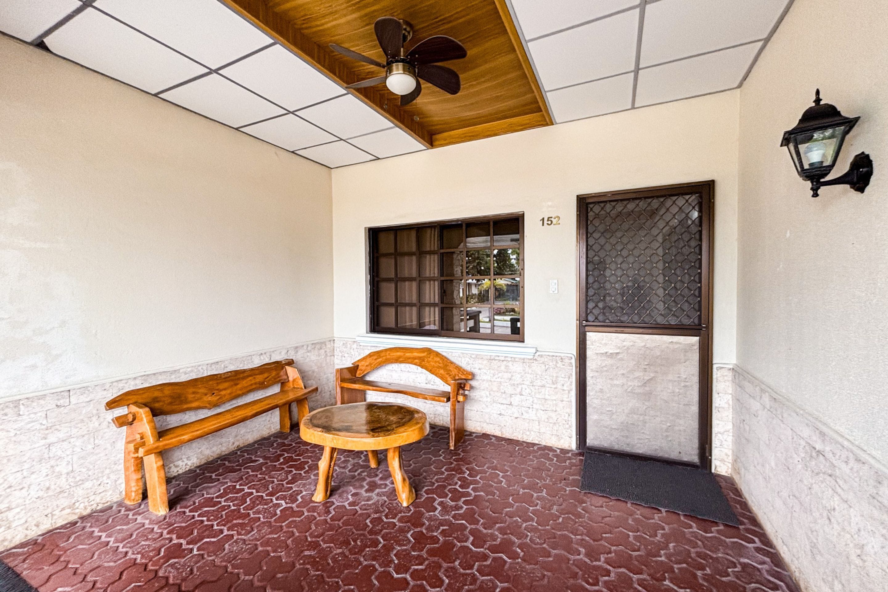 A covered outdoor seating area with a wooden bench and table, a ceiling fan, a window with a view, and a door with a number.