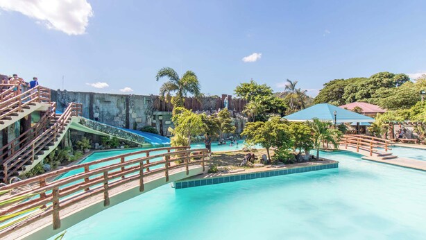 A swimming pool area with wooden bridges and steps leading to the water, surrounded by greenery and a clear sky.