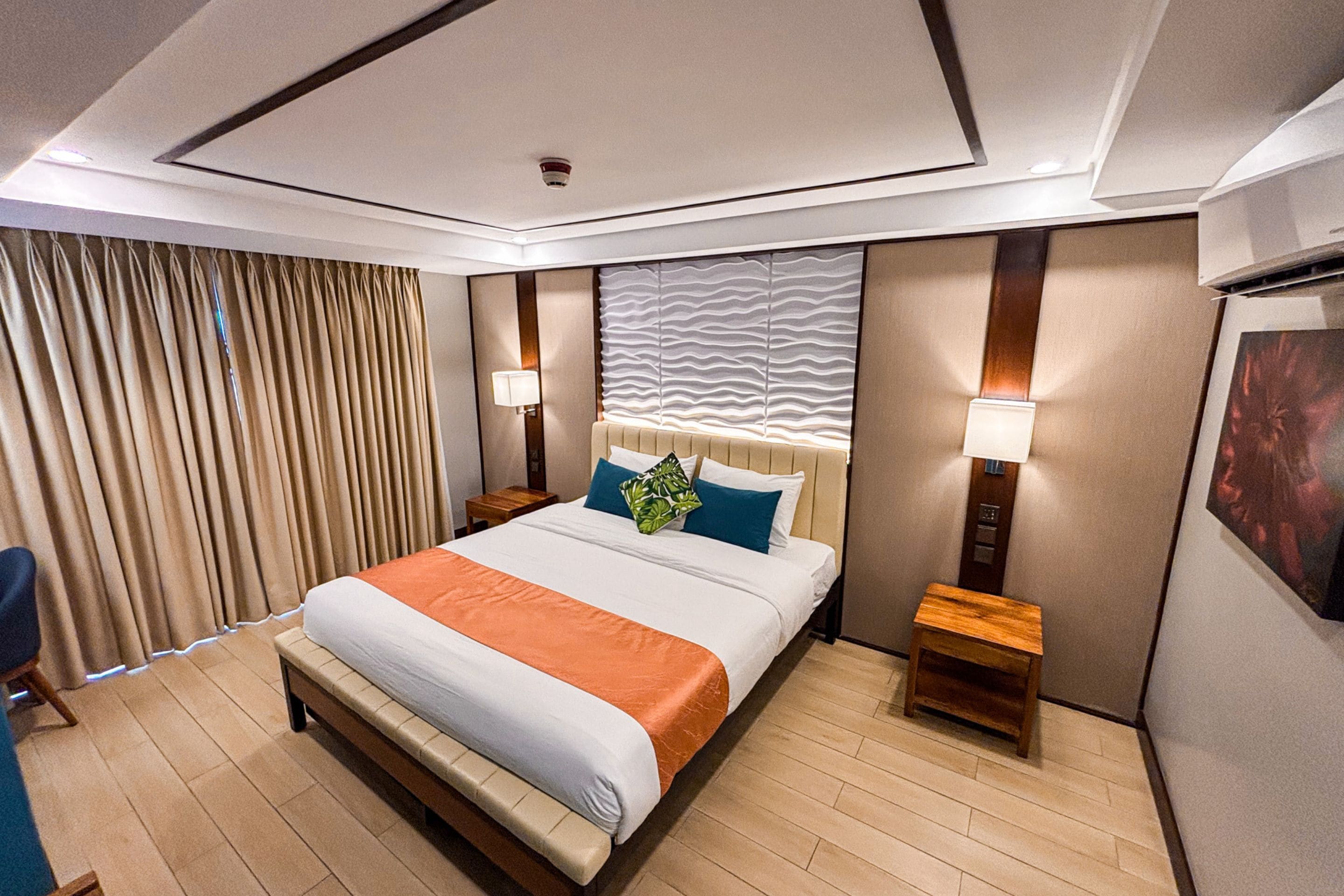 Habitación de hotel con una cama grande, dos mesitas de noche, cabecera con diseño texturizado, lámpara de pared y ventana con cortinas.