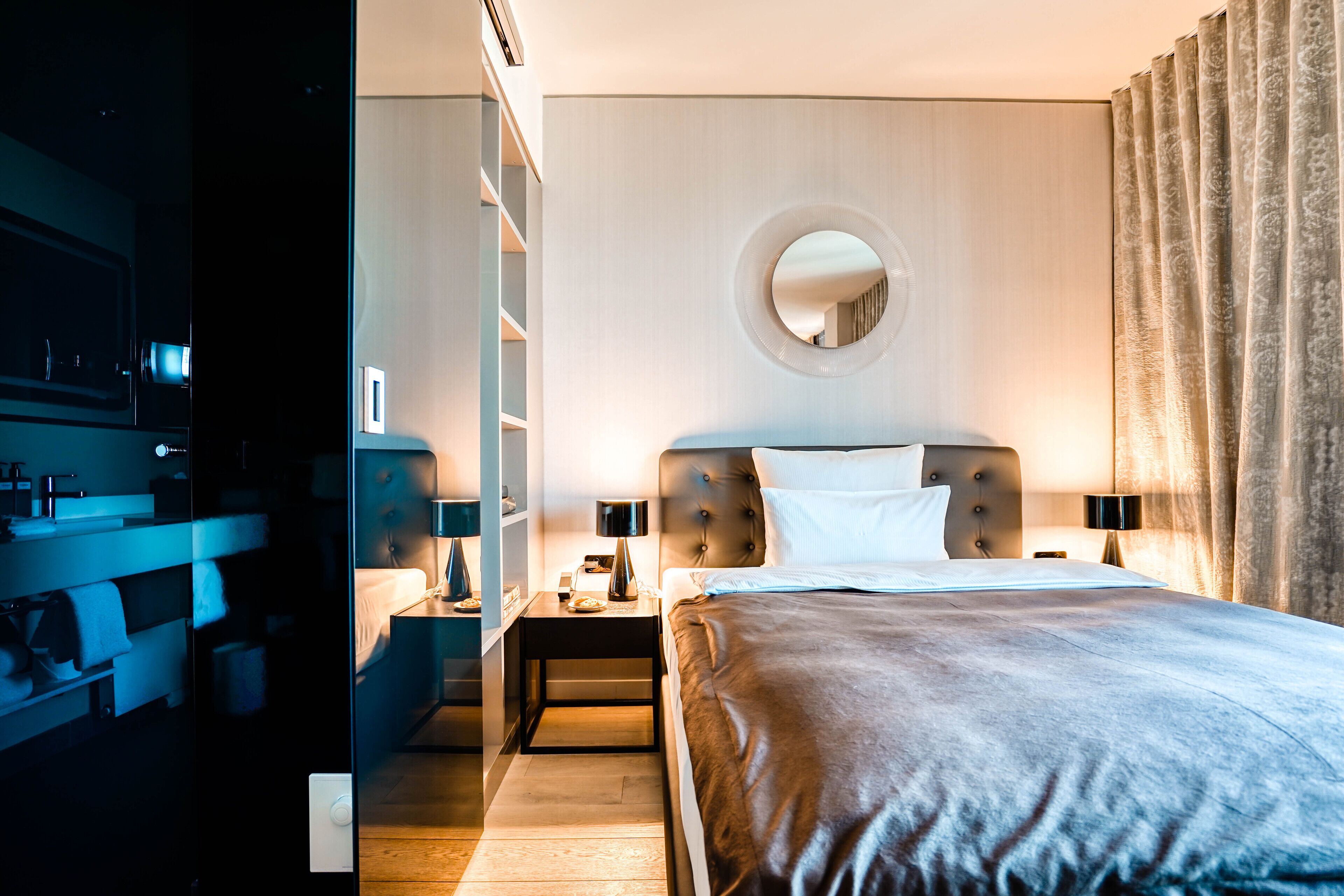 A modern hotel room with a large bed, a round mirror on the wall, a nightstand, and a wardrobe.