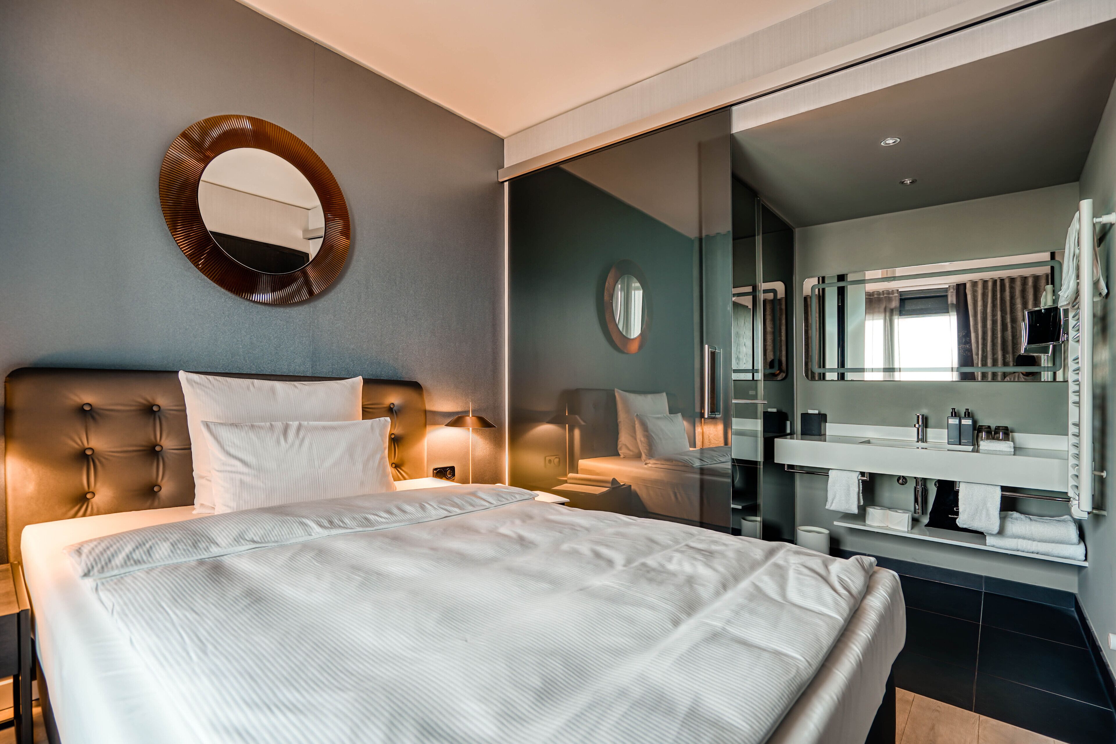 Una habitación de hotel moderna con una cama grande, un baño separado con mampara de vidrio y un espejo redondo en la pared.