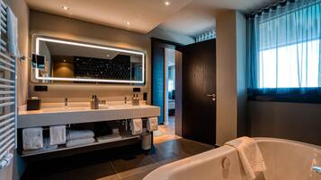 A modern bathroom with a large mirror, two sinks, a bathtub, and a window with curtains.