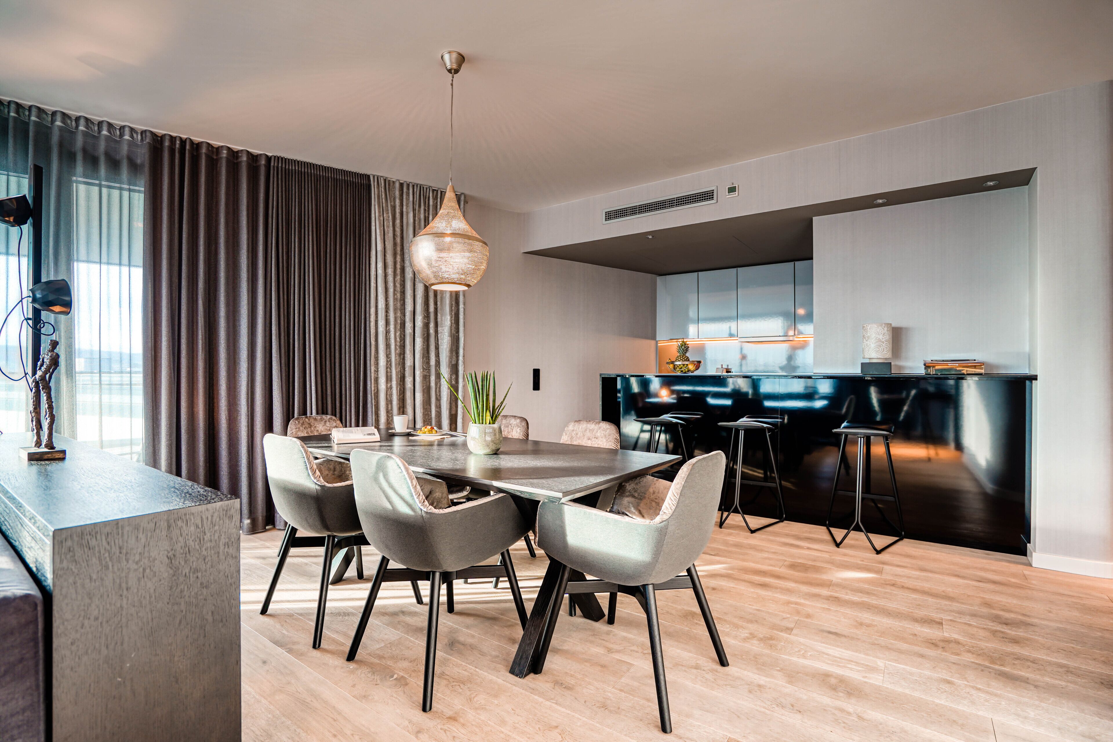 A modern dining area with a wooden floor, a central table with chairs, a pendant light, and a kitchen area with a dark countertop and stools.