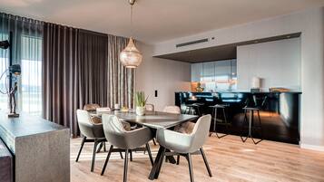 A modern dining area with a wooden floor, a central table with chairs, a pendant light, and a kitchen area with a dark countertop and stools.