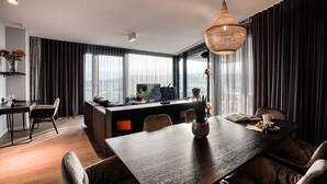 A modern hotel room with a dark wood dining table, comfortable seating, a large window with curtains, and a city view. - CLOUD No7 Apartments (Stuttgart)