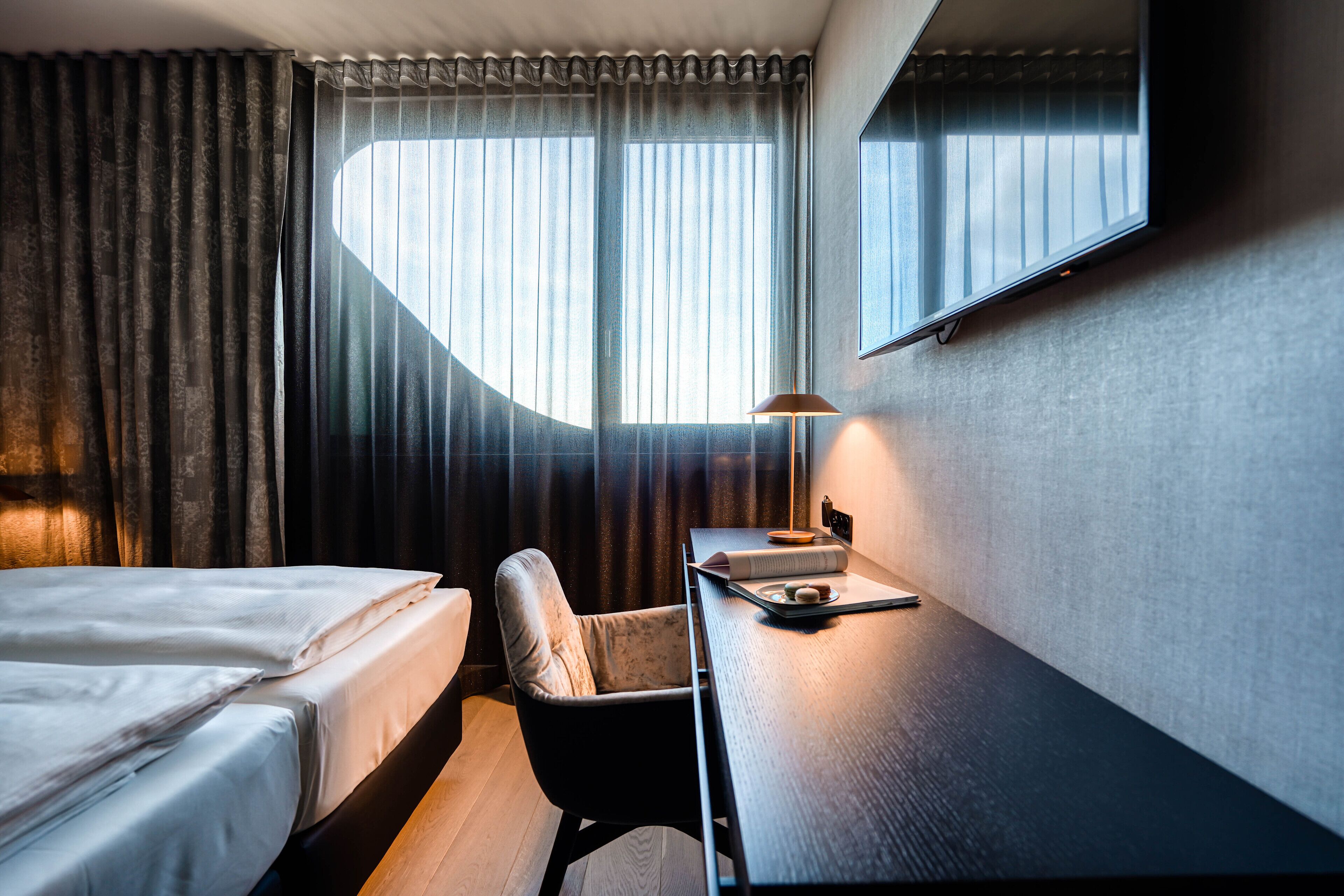 Una camera d'albergo con un letto, una scrivania, una sedia, una lampada e un'ampia finestra con tende.