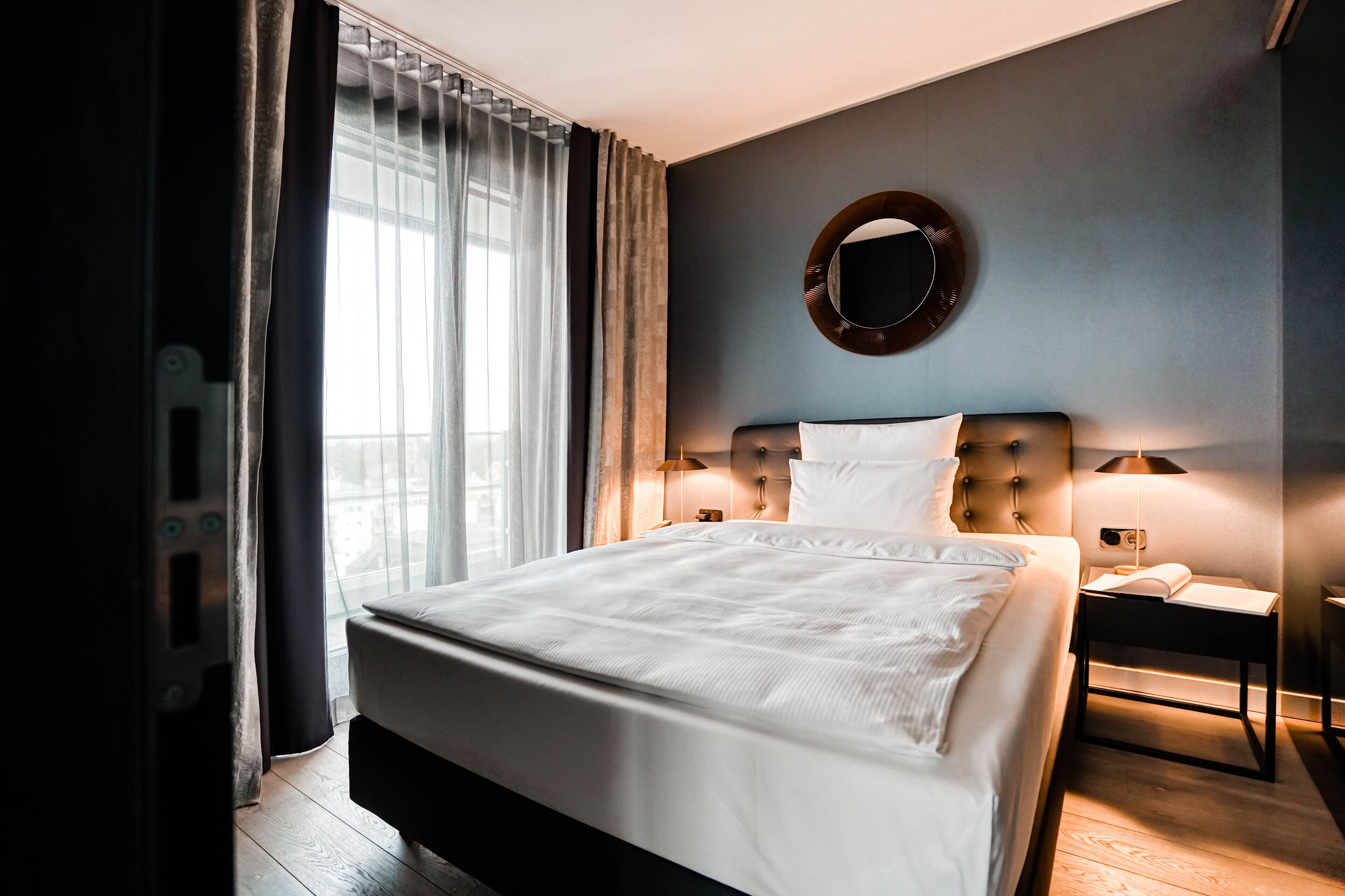 Ein ordentlich bezogenes Bett in einem Hotelzimmer, mit einem runden Spiegel an der Wand, einem Nachttisch mit Lampe und einem Fenster mit Vorhängen.
