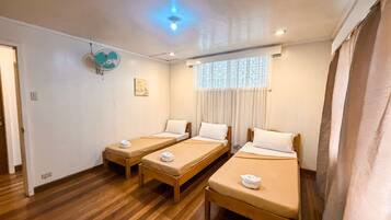 Habitación de hotel con tres camas individuales, ventilador de techo, ventana con cortinas y piso de madera.
