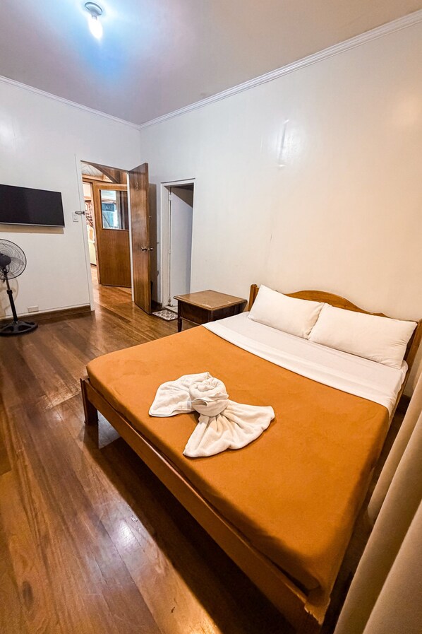 Quarto de hotel com cama, mesa de cabeceira, ventilador e televisão.