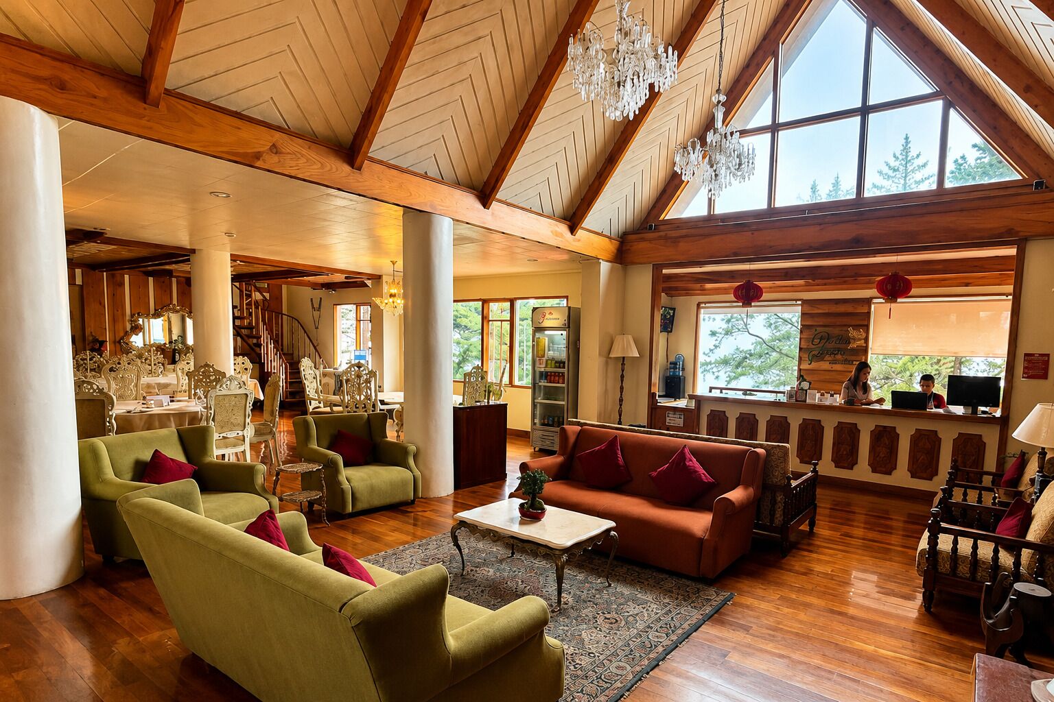 A spacious living area with a high ceiling, wooden beams, and large windows. There are multiple seating arrangements including sofas and chairs.