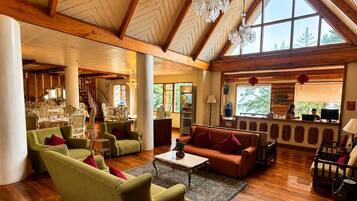 A spacious living area with a high ceiling, wooden beams, and large windows. There are multiple seating arrangements including sofas and chairs.