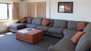 A living room with a grey sectional sofa, orange patterned ottoman, and a framed picture on the wall.