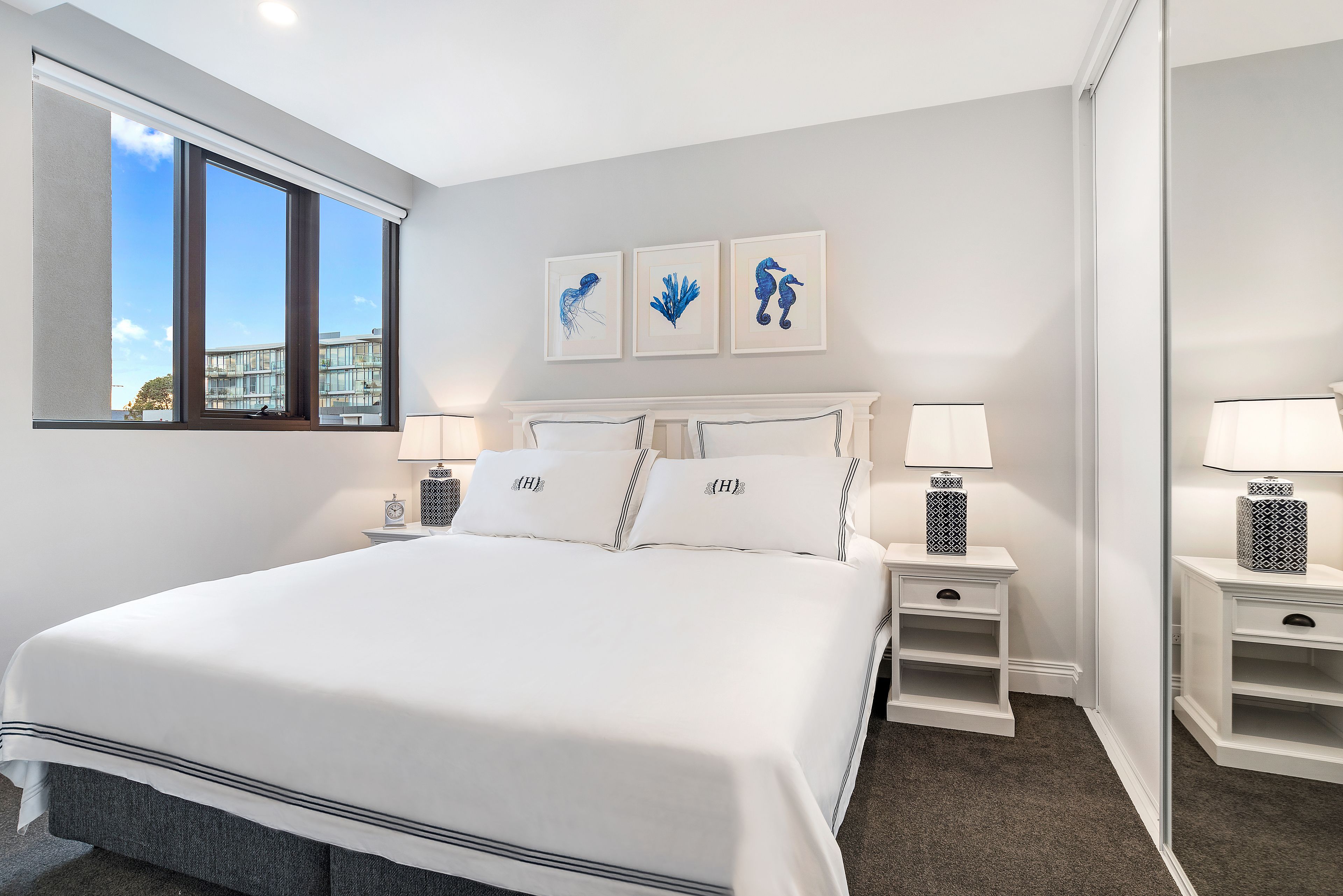 Quarto moderno com uma cama grande, mesas de cabeceira com abajures e vista para um edifício pela janela.