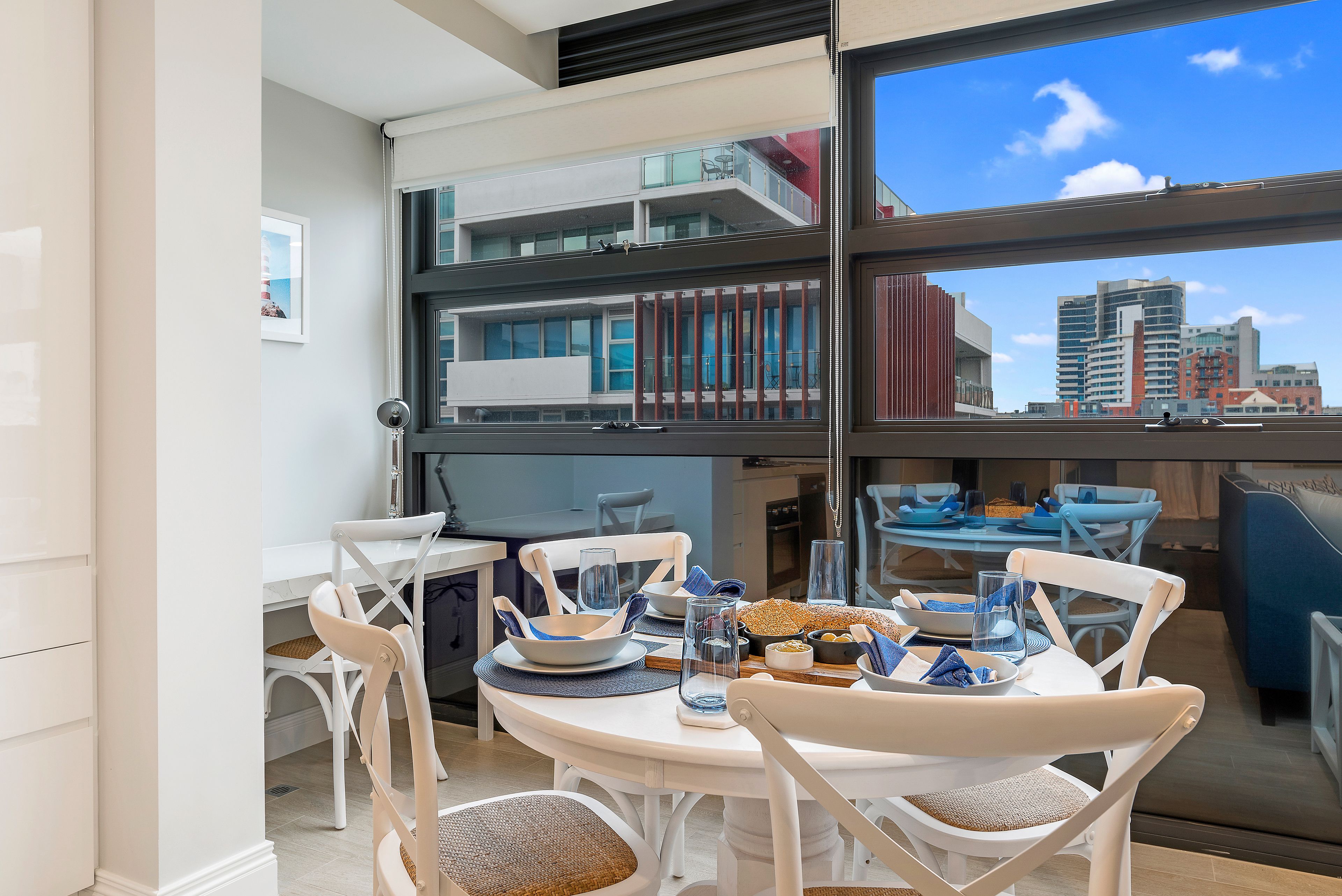 A dining area with a table set for four, white chairs, and a view of a cityscape through large windows.