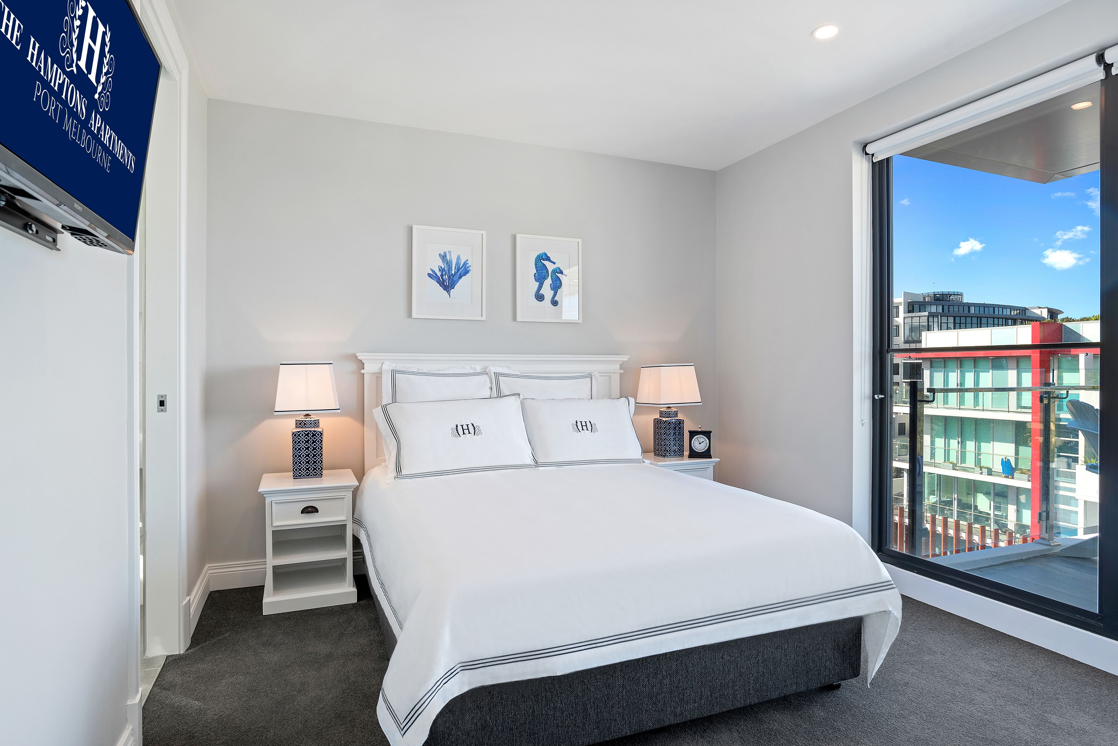 Une chambre d’hôtel moderne avec un grand lit, deux tables de chevet avec des lampes, une vue sur la ville par une grande fenêtre et deux œuvres d’art encadrées accrochées au mur.