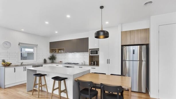 A modern kitchen with white cabinetry, a central island with stools, stainless steel appliances, and a wooden dining table. - Coastal Vibe Apartments (Warrnambool)