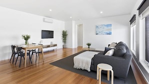 A modern living room with a grey sofa, a wooden dining table with chairs, a flat-screen TV, and a painting on the wall. - Coastal Vibe Apartments (Warrnambool)