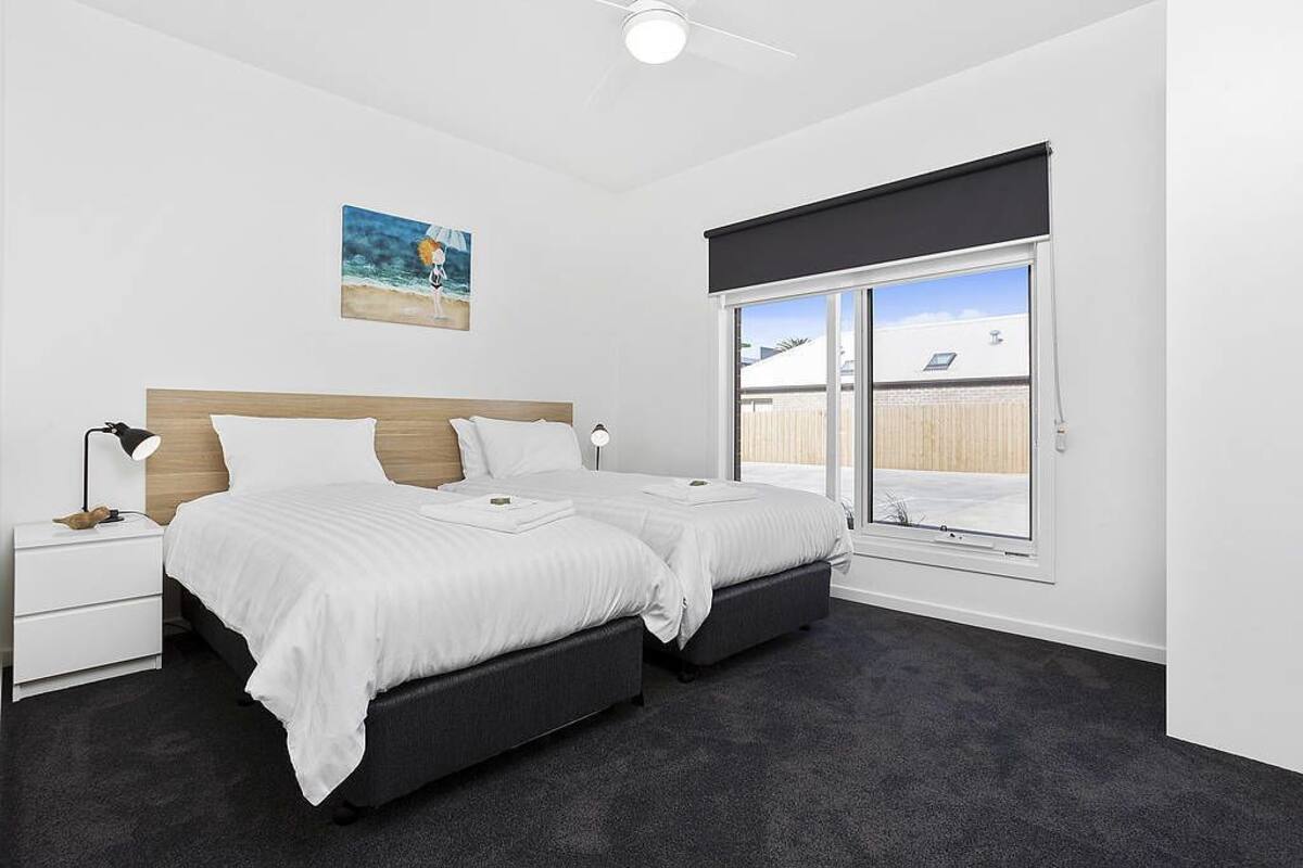 A bedroom with two beds, a nightstand, a ceiling fan, a window with a view, and a painting on the wall.