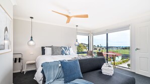 A modern bedroom with a bed, a grey sofa, a ceiling fan, and a large window offering a view of a green landscape. - Madsen Retreat (Penguin)