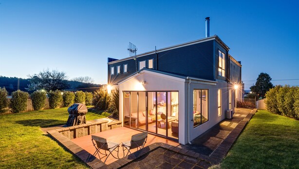 A modern house with a large glass door leading to a patio area with outdoor furniture. - Madsen Retreat (Penguin)