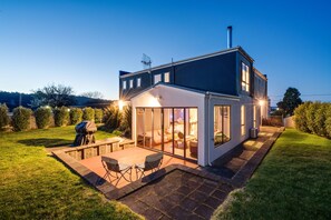 A modern house with a large glass door leading to a patio area with outdoor furniture. - Madsen Retreat (Penguin)