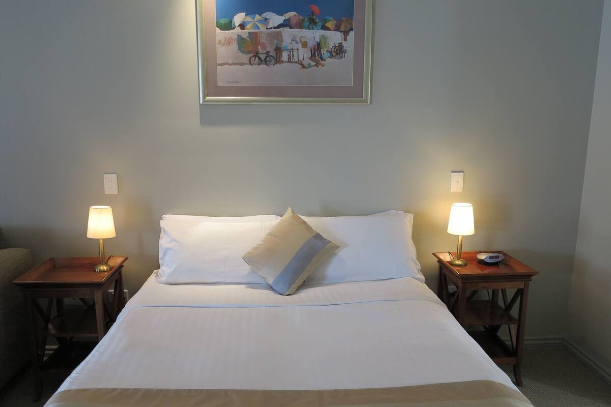 A hotel room with a bed, two bedside tables with lamps, a framed painting on the wall, and a chair in the corner.