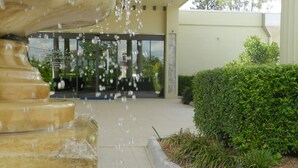 A fountain with water spouting from its top, a paved pathway leading to a building entrance, and a well-maintained hedge beside the pathway. - Shangri La Gardens (Wynnum West)