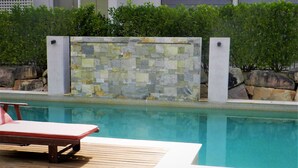 A swimming pool with a tiled wall and a wooden deck with a lounge chair. - Shangri La Gardens (Wynnum West)