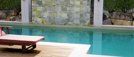 Una piscina con un muro de azulejos y una terraza de madera con una tumbona.