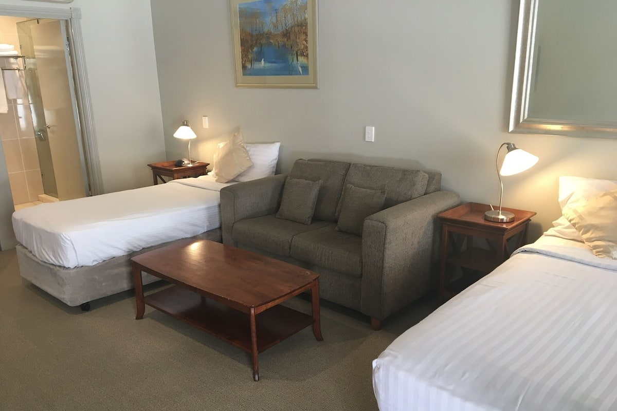 A hotel room with two single beds, a sofa, a wooden coffee table, and two bedside lamps.
