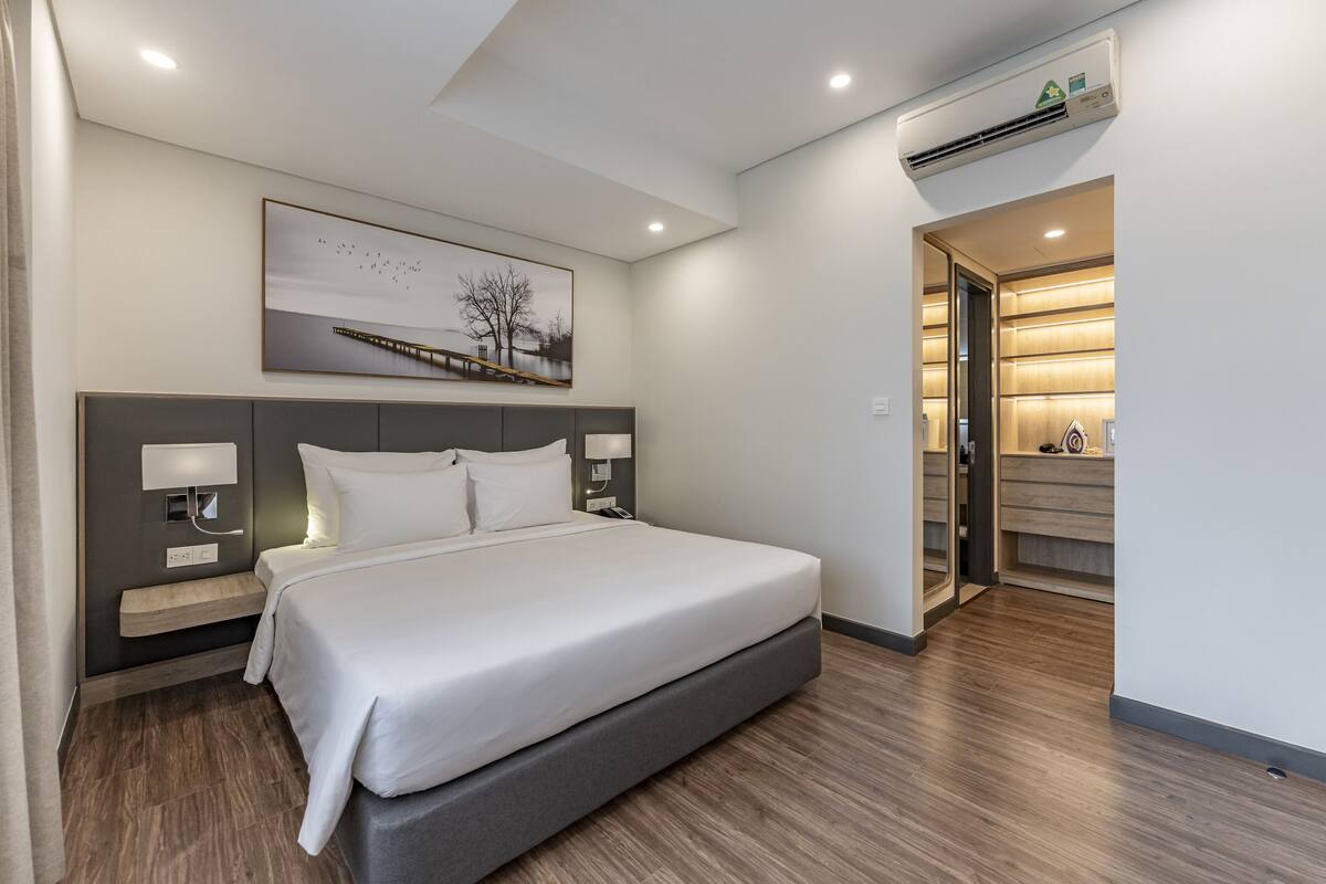 Une chambre d’hôtel moderne avec un grand lit, des tables de chevet, une petite table, un appareil de climatisation et un tableau encadré au mur.