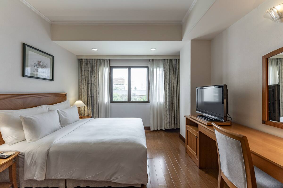 Une chambre d’hôtel avec un grand lit, un téléviseur posé sur un meuble en bois, un bureau et une fenêtre avec des rideaux.