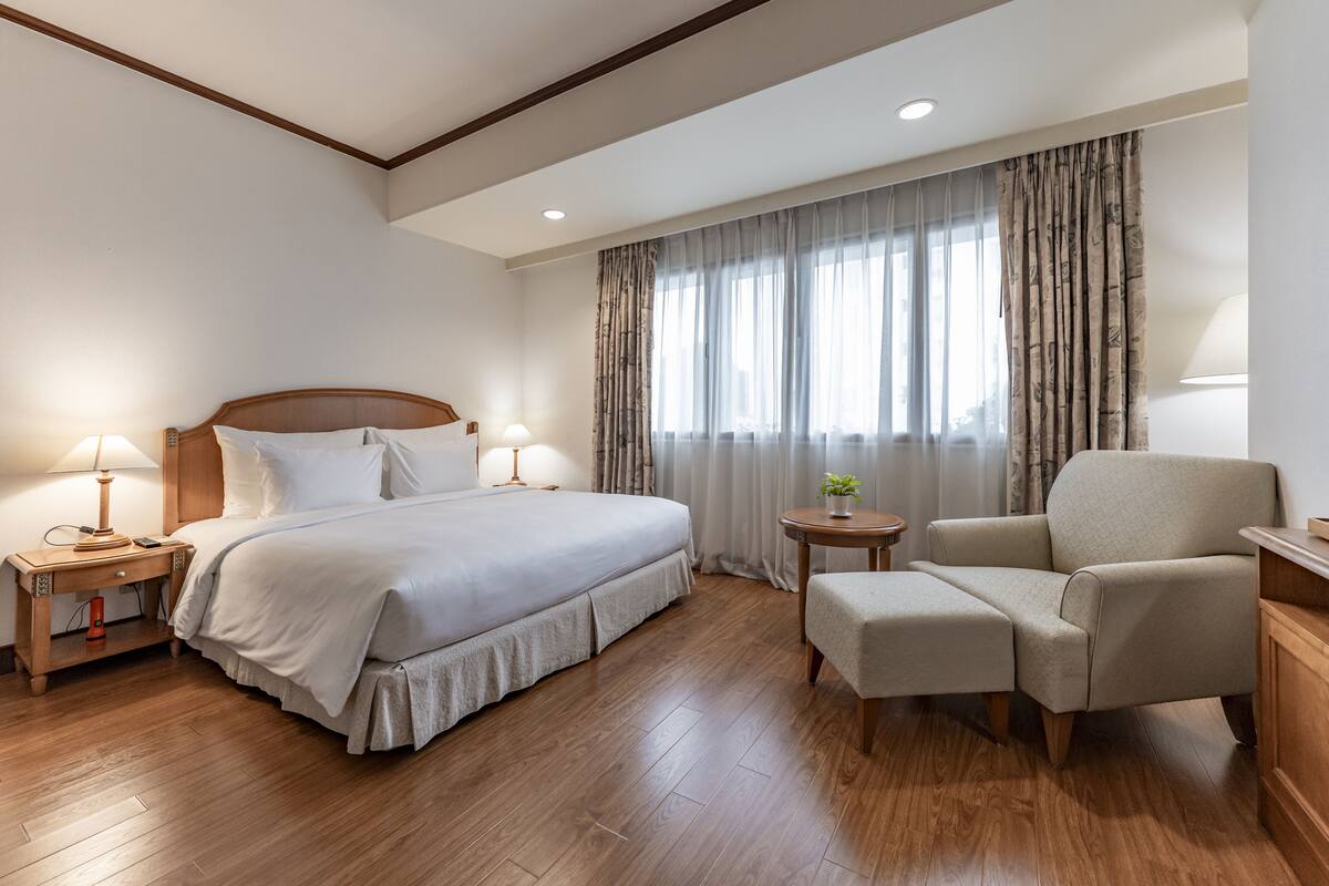 Une chambre d’hôtel avec un grand lit, des tables de chevet avec des lampes, une chaise, une petite table avec une plante et une fenêtre avec des rideaux.