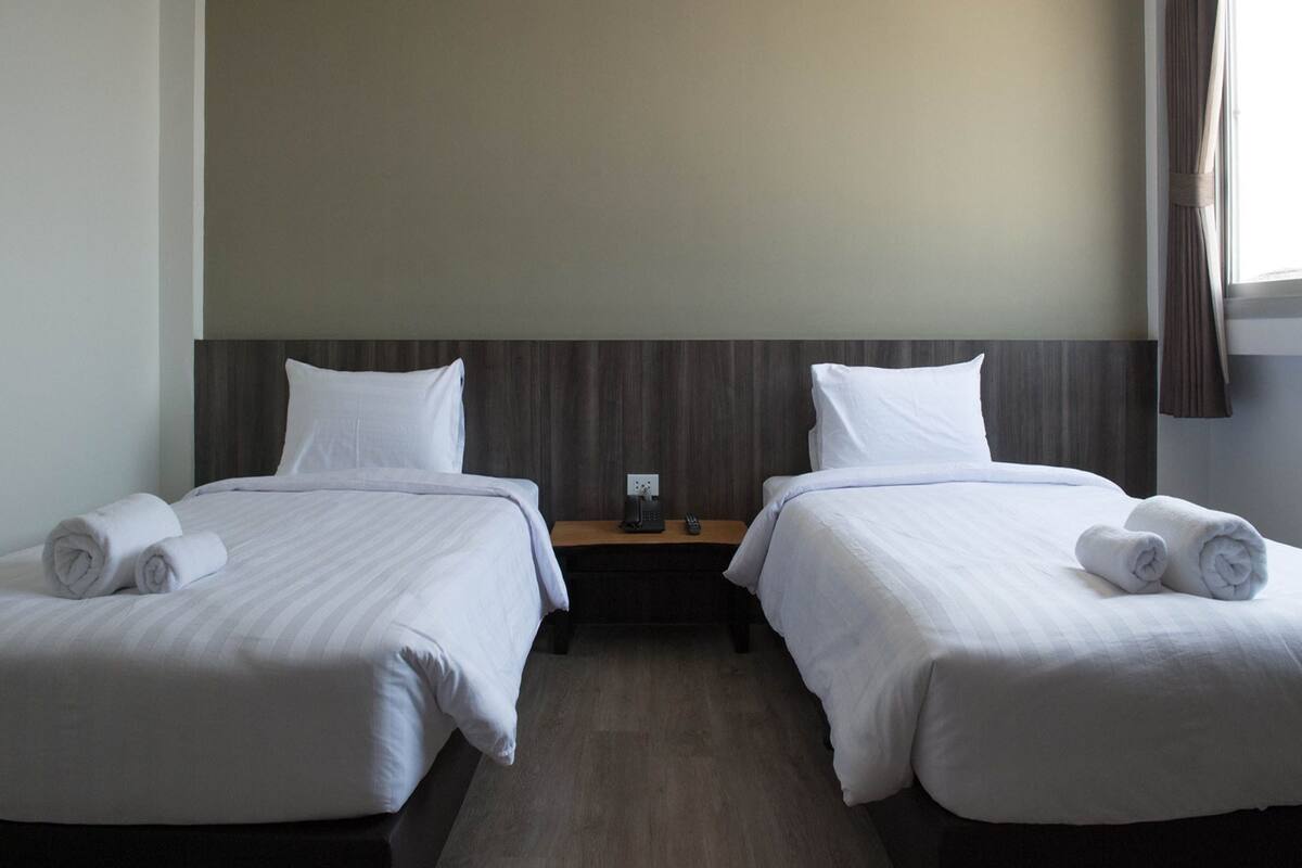 Une chambre d’hôtel avec deux lits, chacun doté de draps et d’oreillers blancs, d’une tête de lit en bois, d’une table de chevet avec une lampe et d’une fenêtre avec des rideaux.