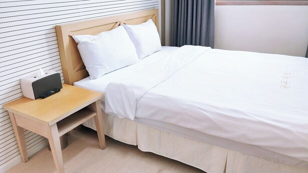 A neatly made bed with white linens, a wooden nightstand with a remote control, and a bedside table.