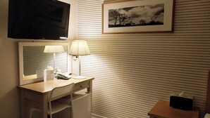 A hotel room with a large mirror, a flat-screen TV, a desk with a lamp, a chair, a bedside table with a phone and a tissue box.